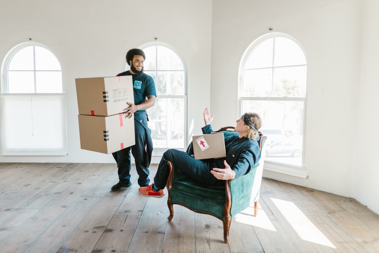 Two movers handling cardboard boxes in a bright room with large windows. Casual and efficient.