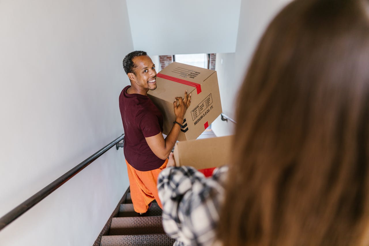 An adult man smiling while carrying a moving box up staircase indoors.
